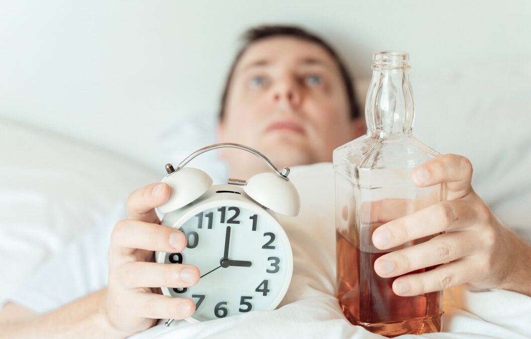 Man laying in bed, wide awake, holding an alarm clock and a bottle of alcohol